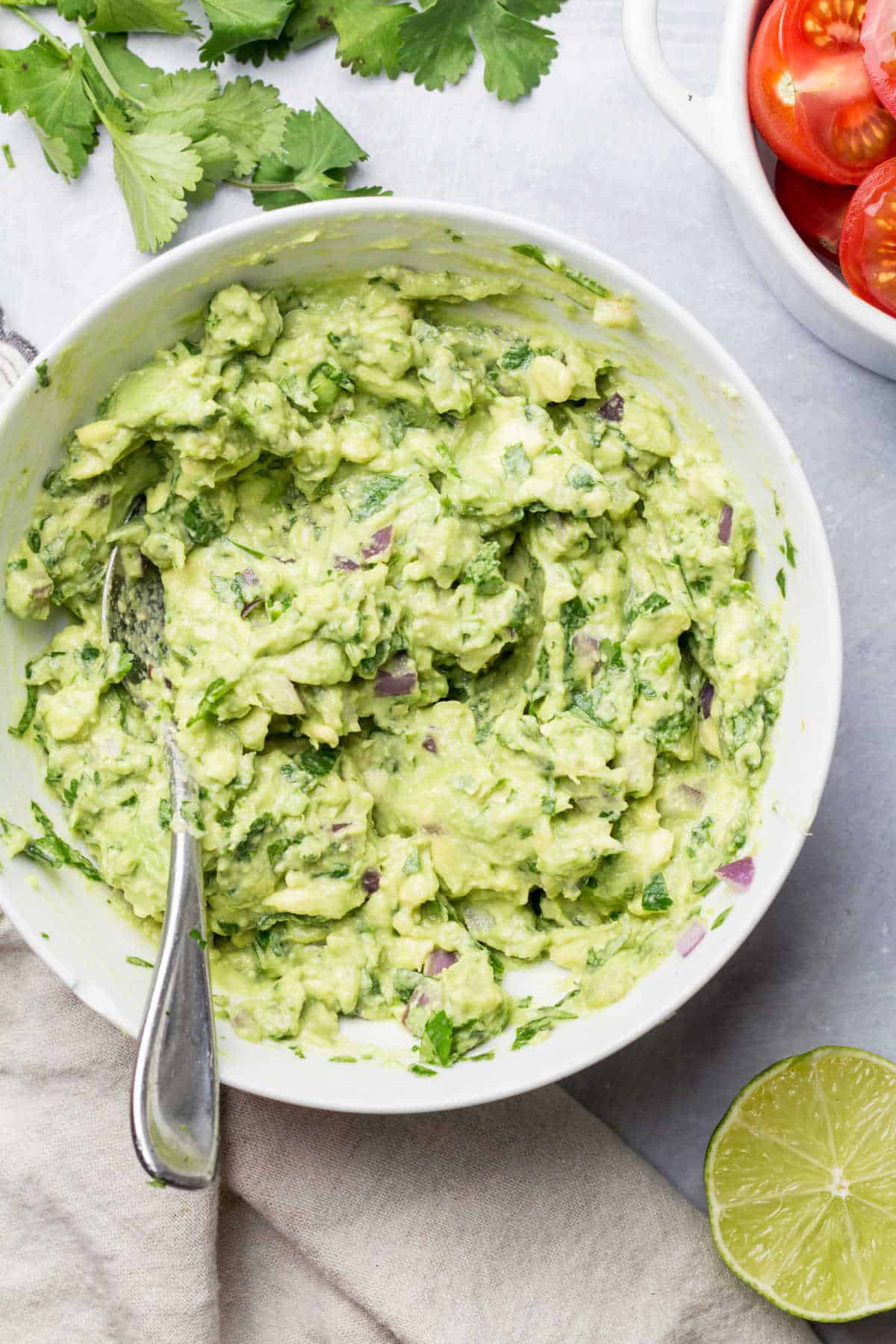 A dish of homemade guacamole next to cilantro tomatoes and lime.