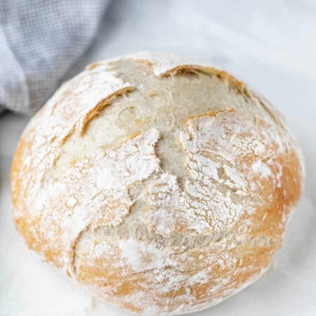 A loaf of no knead bread next to a blue checkered linen.