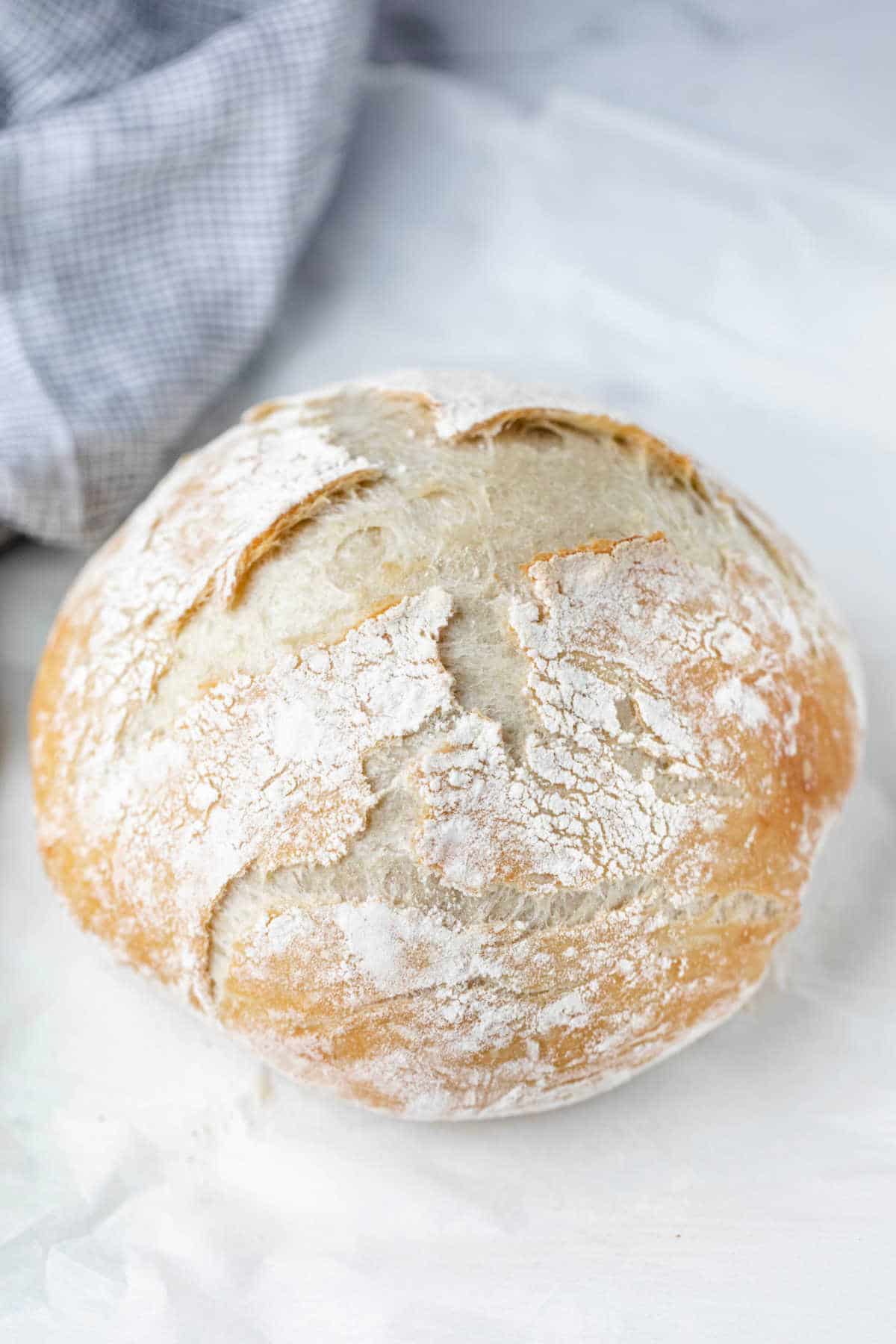 A loaf of no knead bread next to a blue checkered linen. 