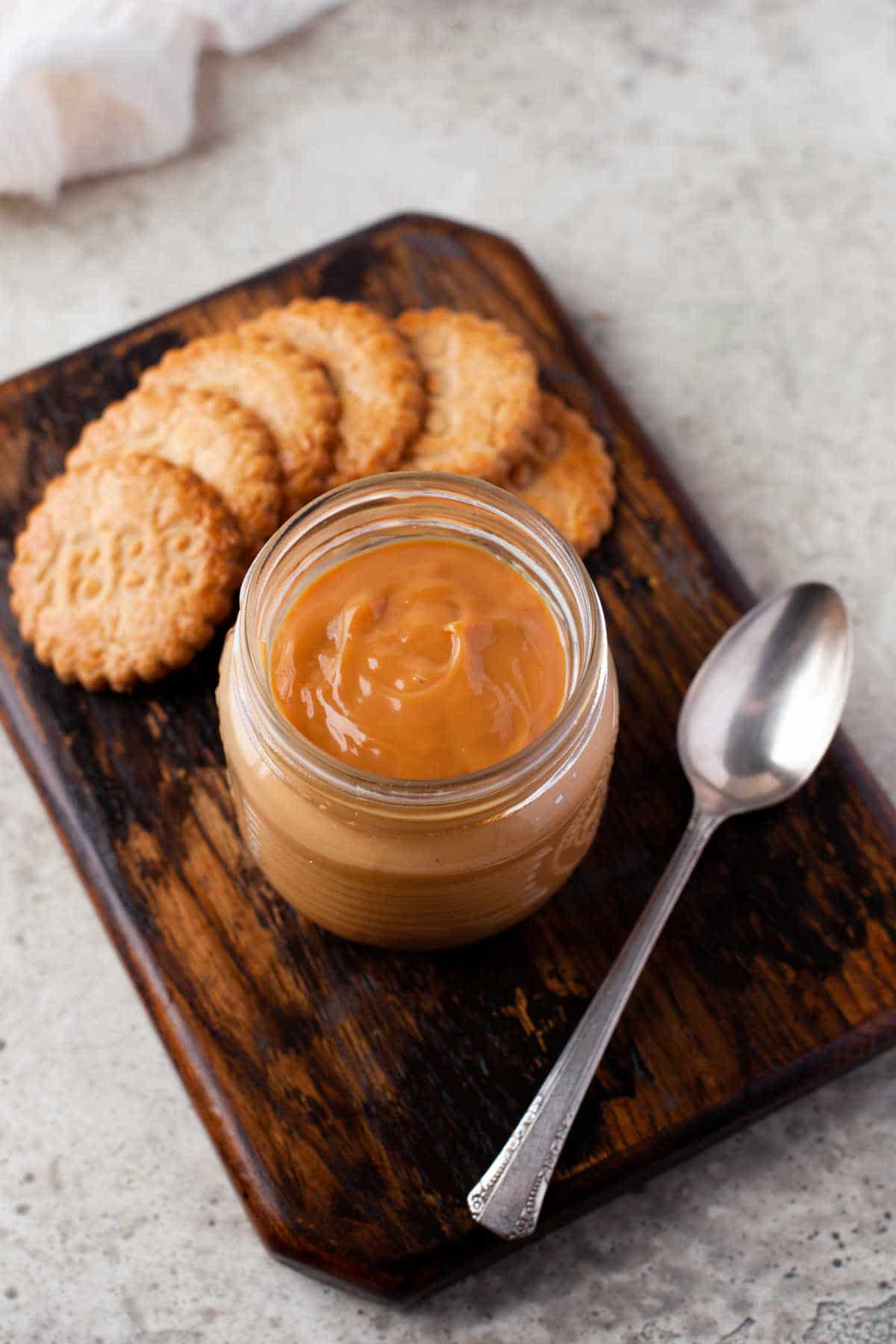 A jar of dulce de leche and a silver spoon on a wooden cutting board. 