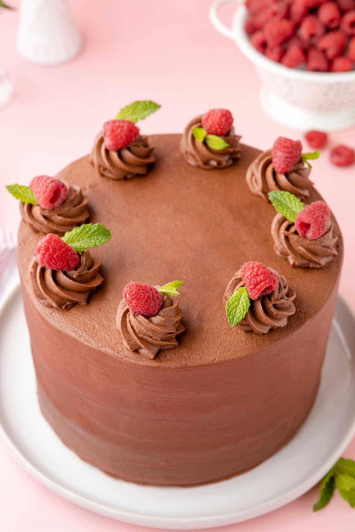 A whole chocolate raspberry cake next to a colander of raspberries. 