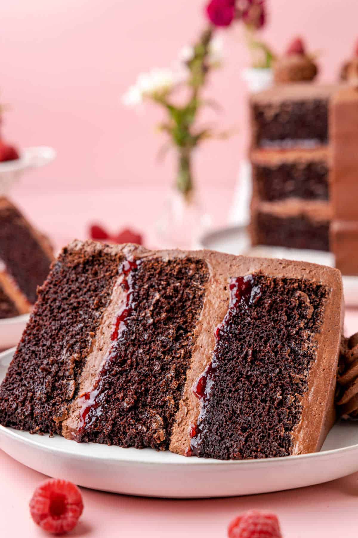 A slice of chocolate raspberry cake on a white plate with two raspberries next to it. 