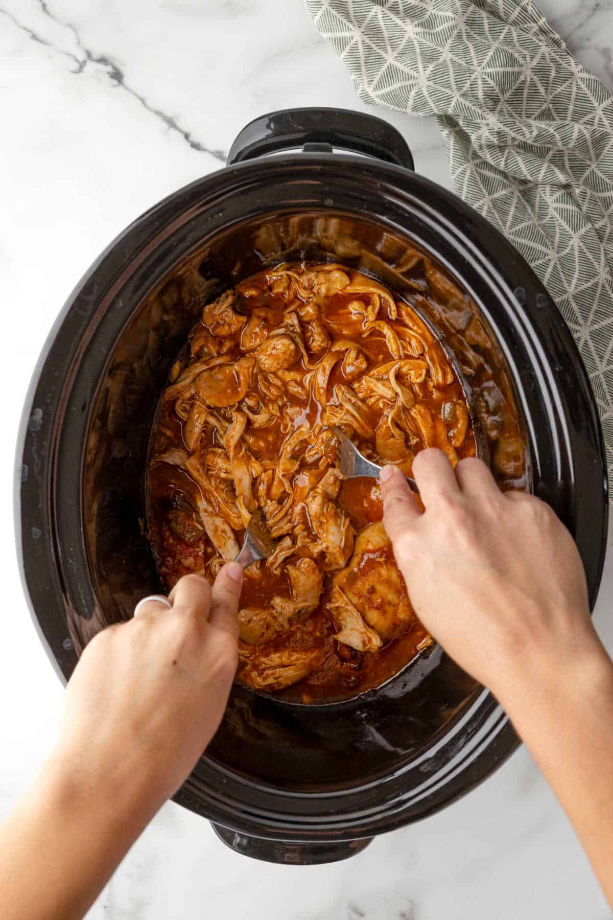 Two hands shredded chicken in a crock pot with forks.
