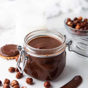 Glass jar of homemade nutella next to hazelnuts and a knife with nutella on it.