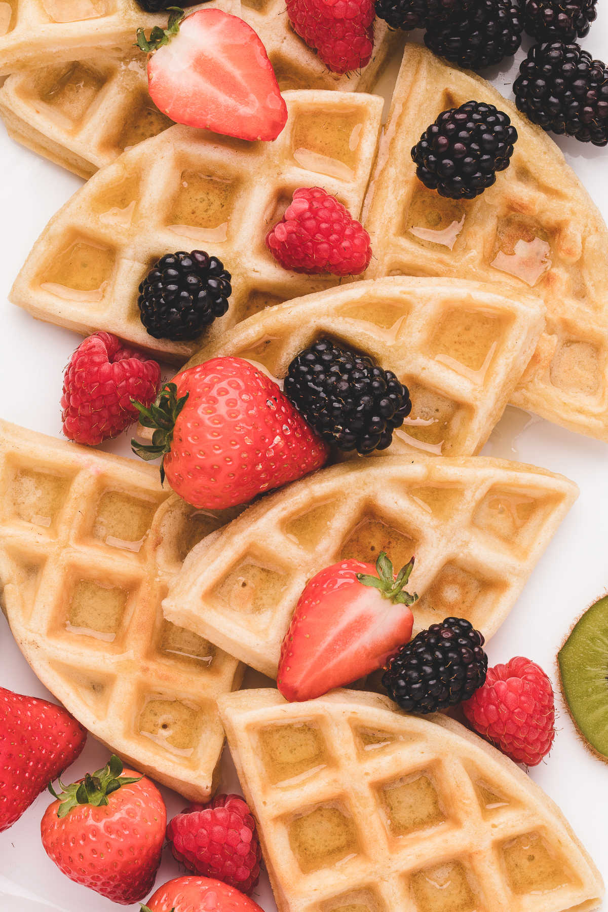 Pieces of Belgian waffle on a white plate topped with syrup and fresh berries. 
