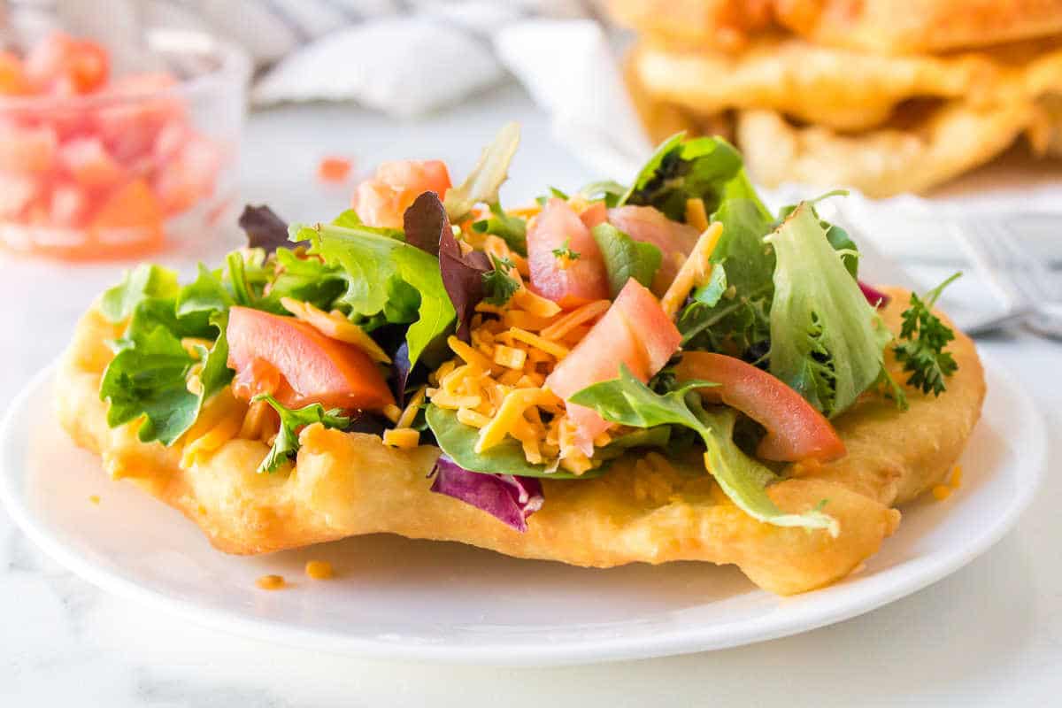 A piece of fry bread with toppings next to a dish of tomatoes.