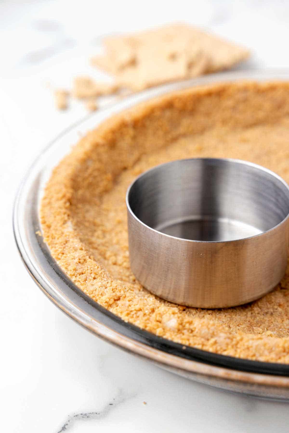 A metal measuring cup pressing graham cracker crumbs into a pie plate. 