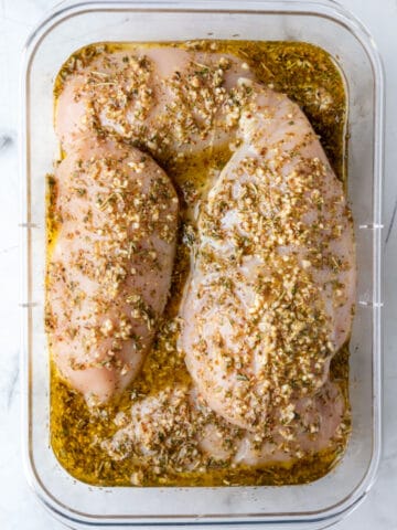 Raw chicken breast in Greek marinade in a container.