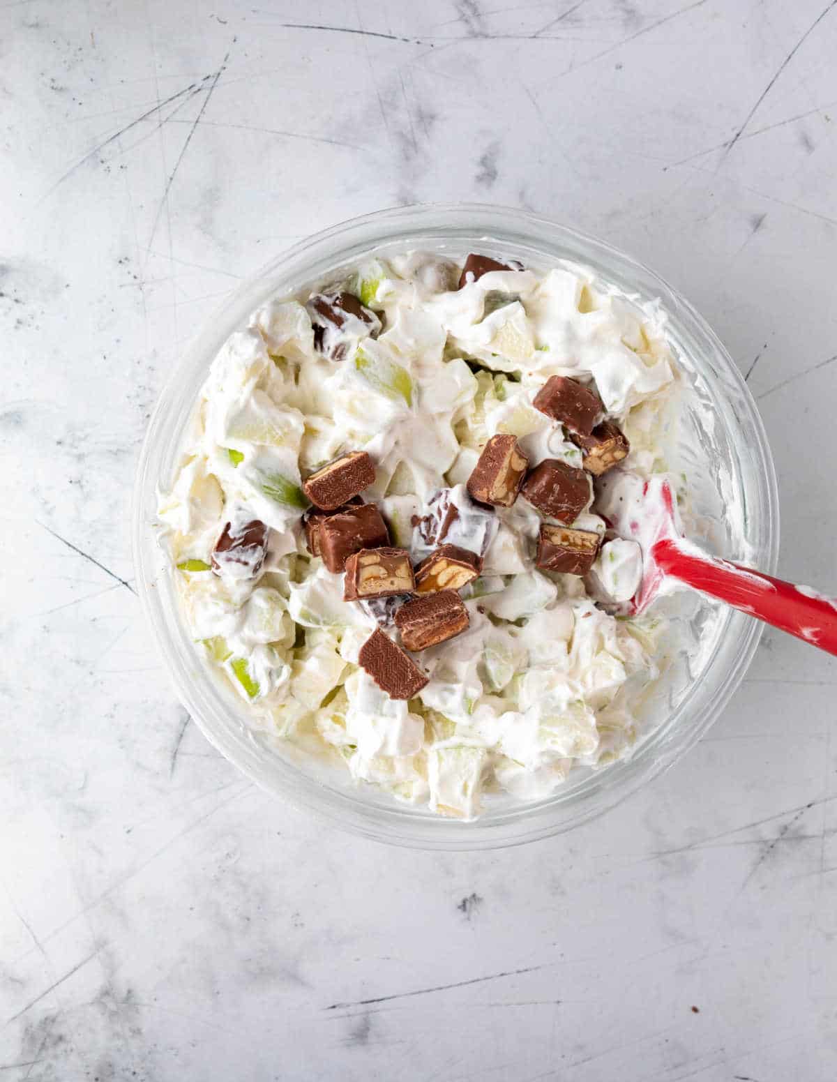 A red rubber spatula folding cool whip and snickers bar pieces into apples. 