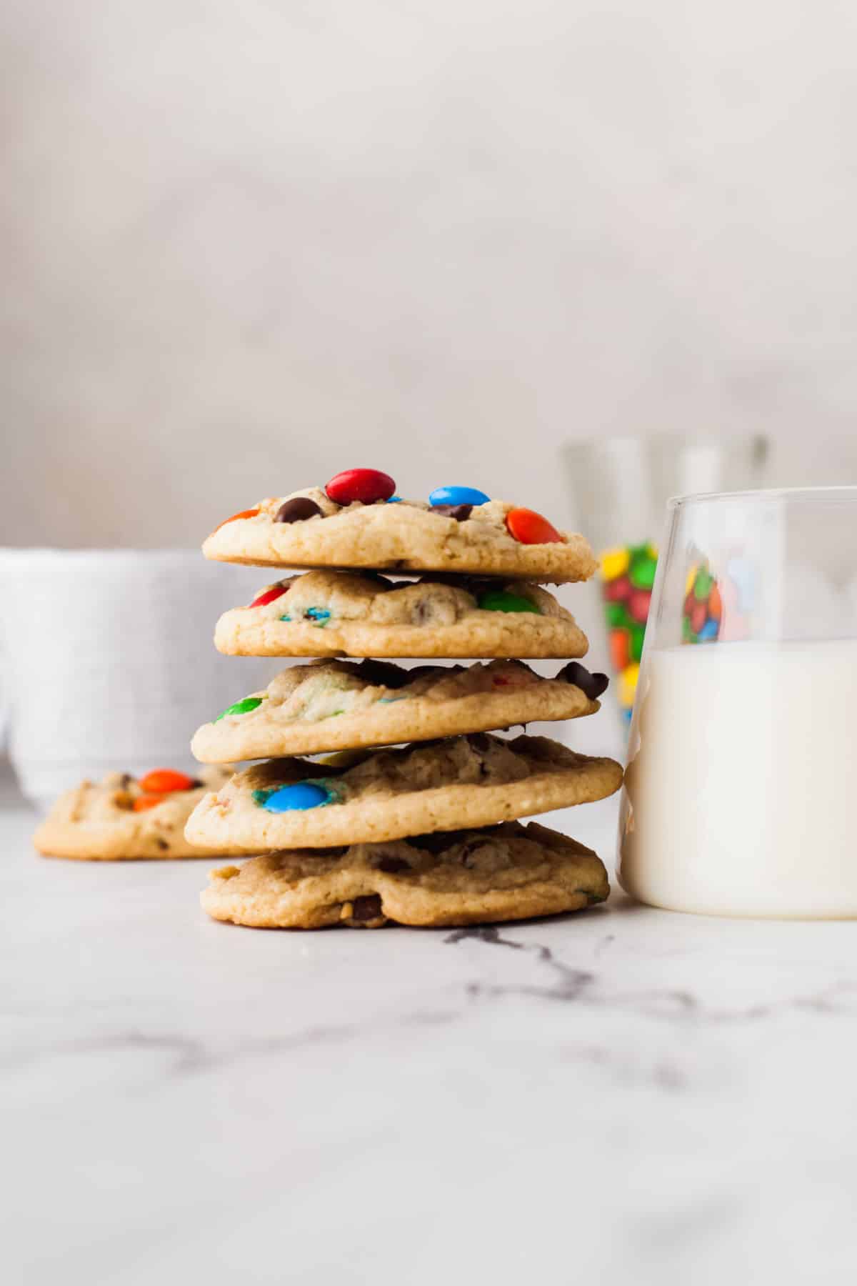 A stack of M&M cookies next to a glass of milk. 