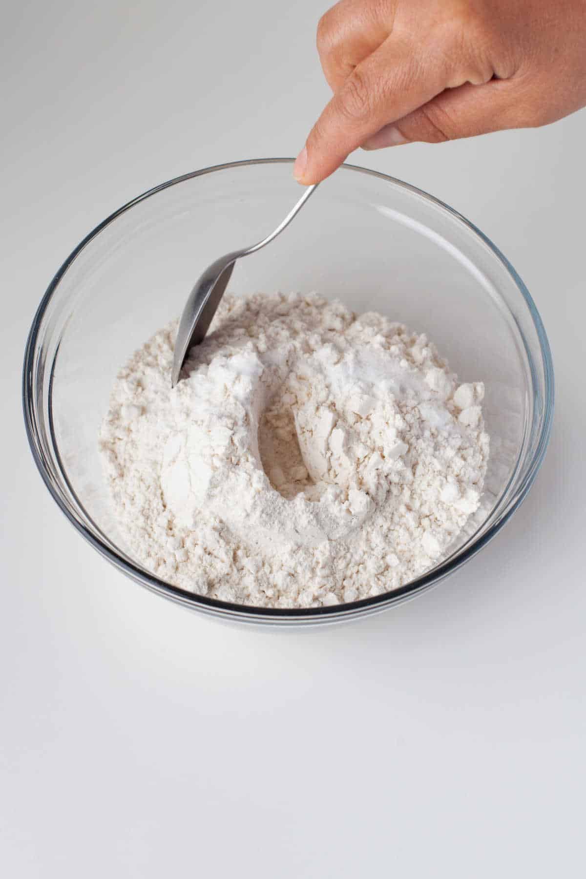 A spoon stirring together flour baking soda and salt in a glass mixing bowl. 