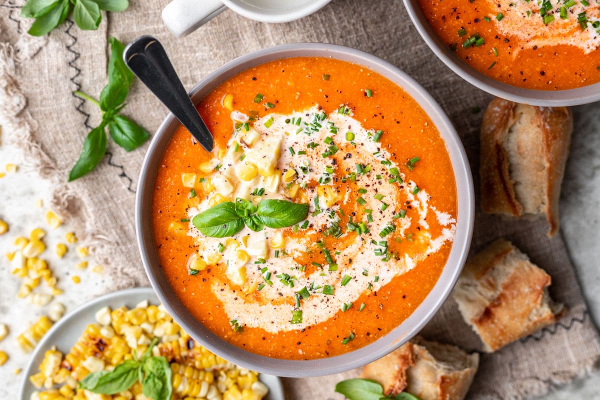A bowl of roasted tomato and corn soup next to bread and a dish of roast corn. 