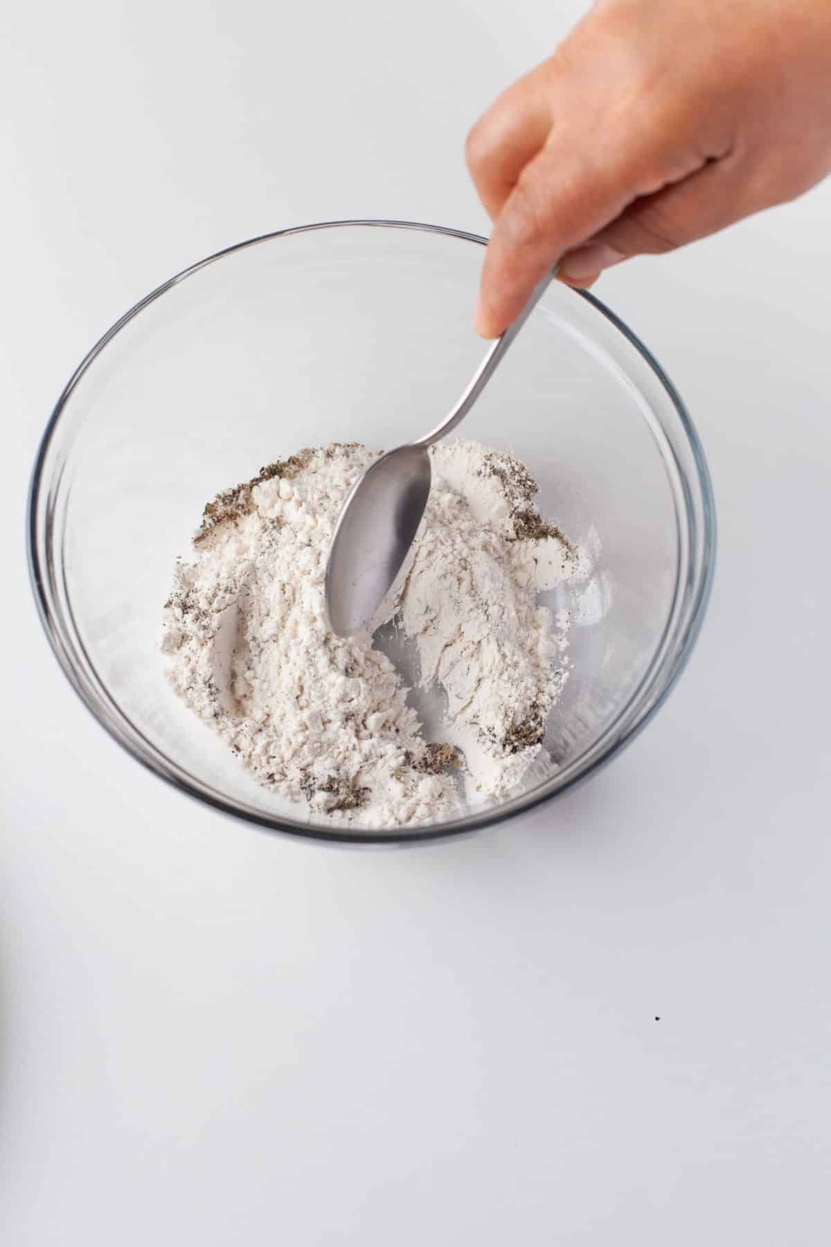 A spoon stirring together flour and seasonings in a glass mixing bowl. 