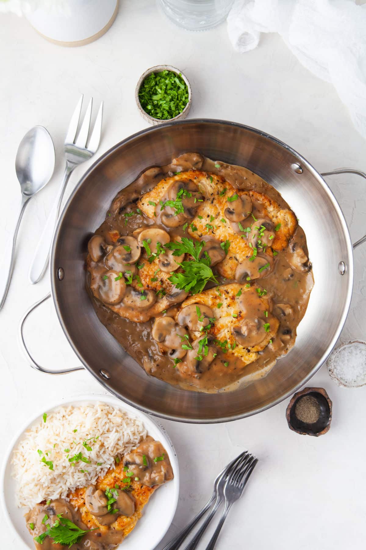 A large silver skillet next to a plate of chicken marsala. 