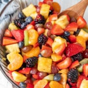 A wooden spoon in a glass mixing bowl full of fruit salad.