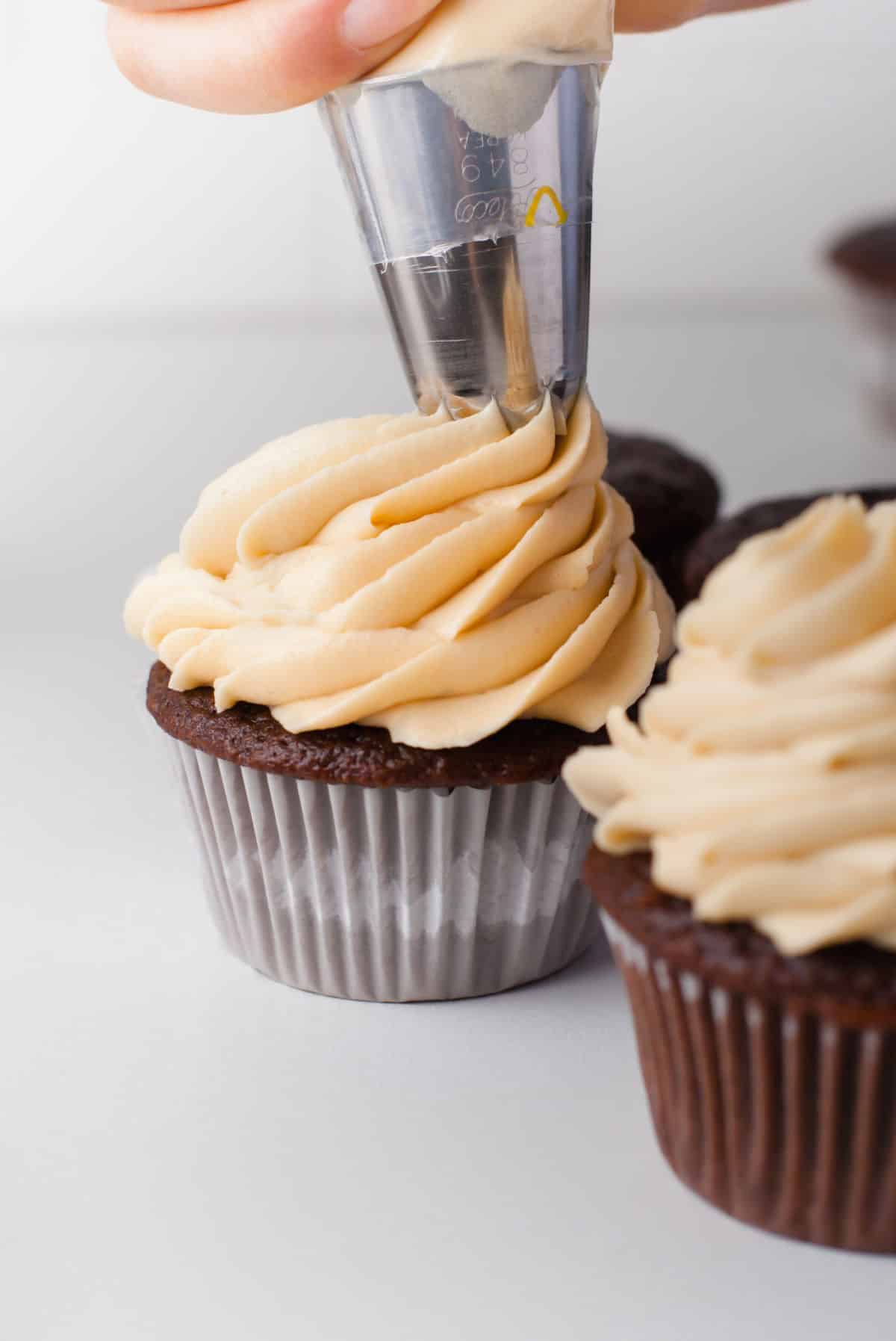 A piping bag piping caramel frosting onto a chocolate cupcake. 