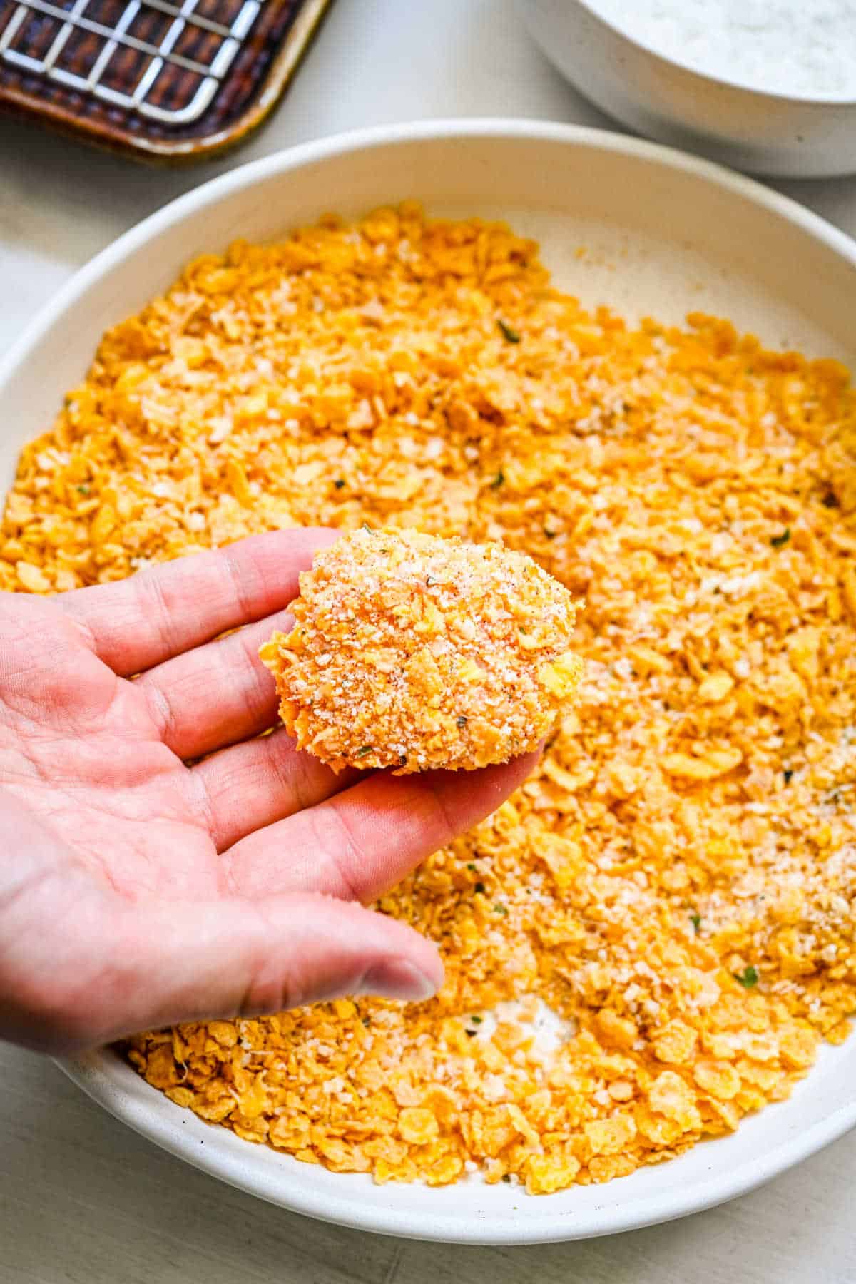 A hand holding a coated chicken nugget above a dish of cornflake seasoning. 