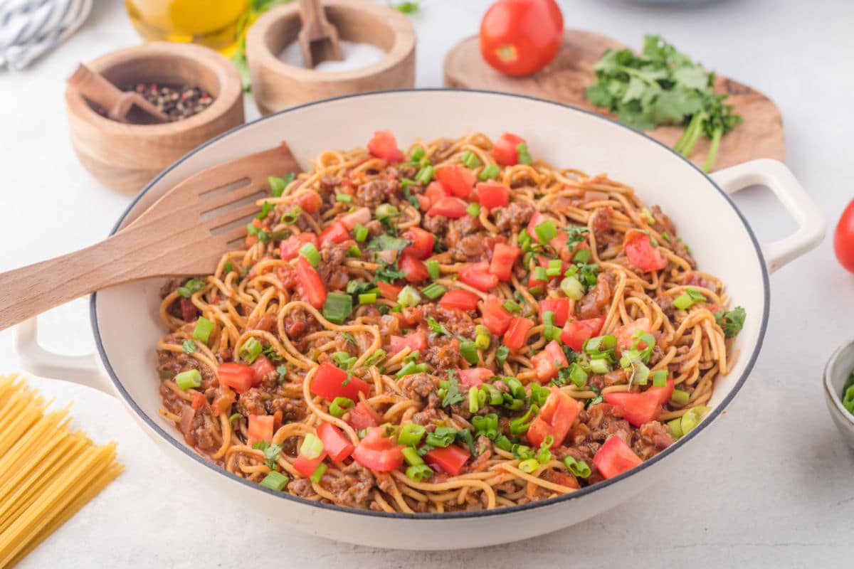 A skillet of taco spaghetti next to taco spaghetti ingredients. 