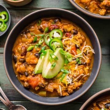 A bowl of turkey pumpkin chili topped with sliced avocado jalapeno and green onion.