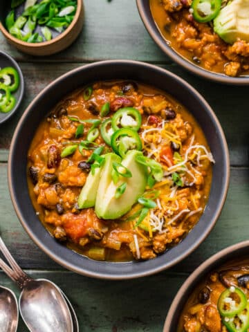 A bowl of turkey pumpkin chili topped with sliced avocado jalapeno and green onion.
