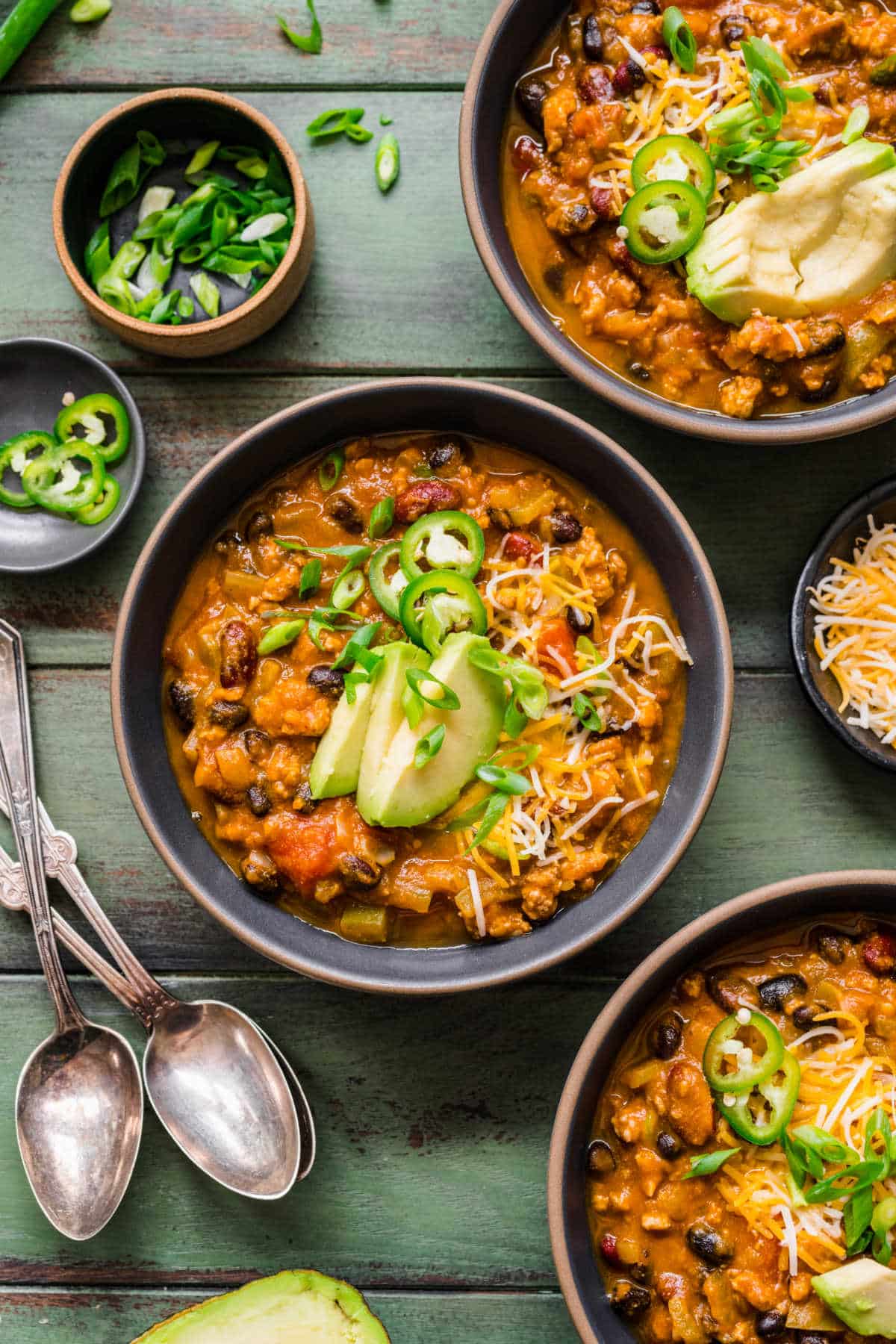 A bowl of pumpkin turkey chili surrounded by dishes of jalapeno and green onion. 