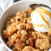 A white dish with apple crisp and a scoop of vanilla ice cream topped with caramel sauce.