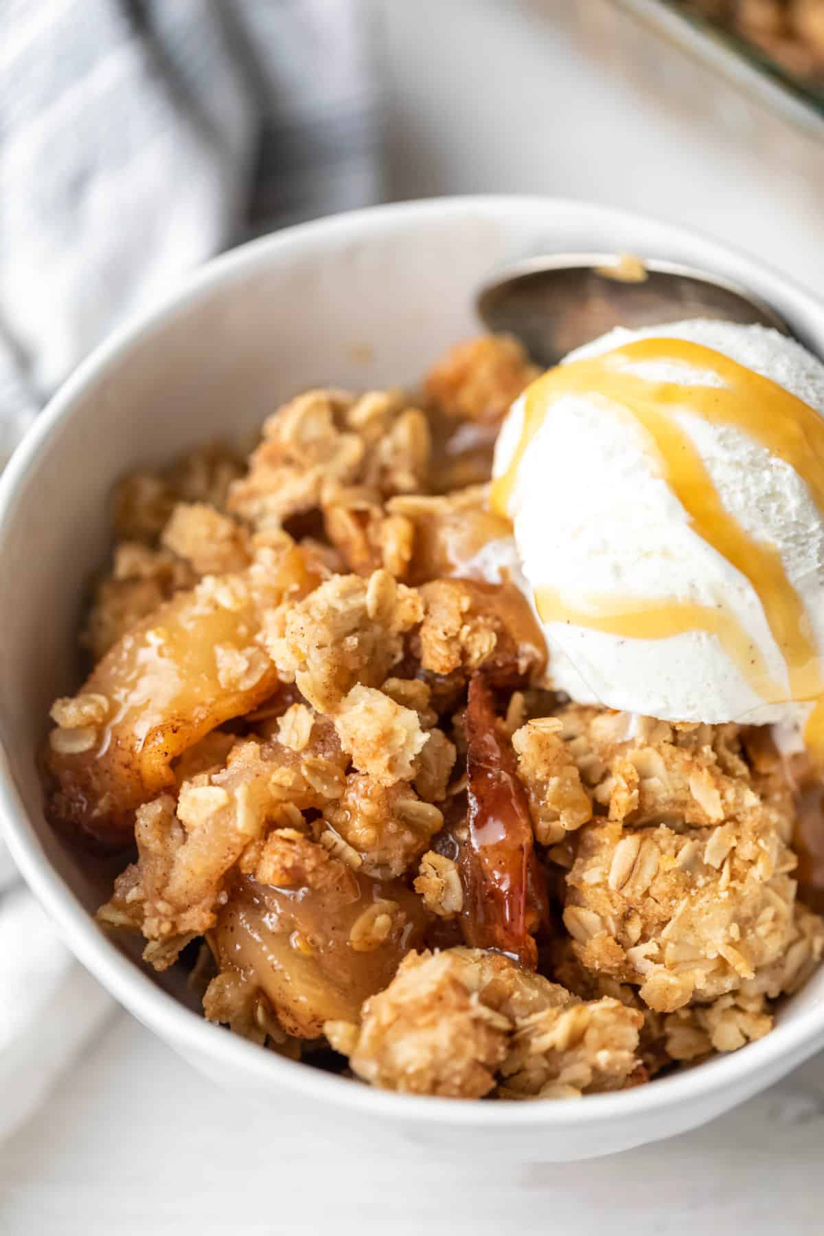 A white dish with apple crisp and a scoop of vanilla ice cream topped with caramel sauce.