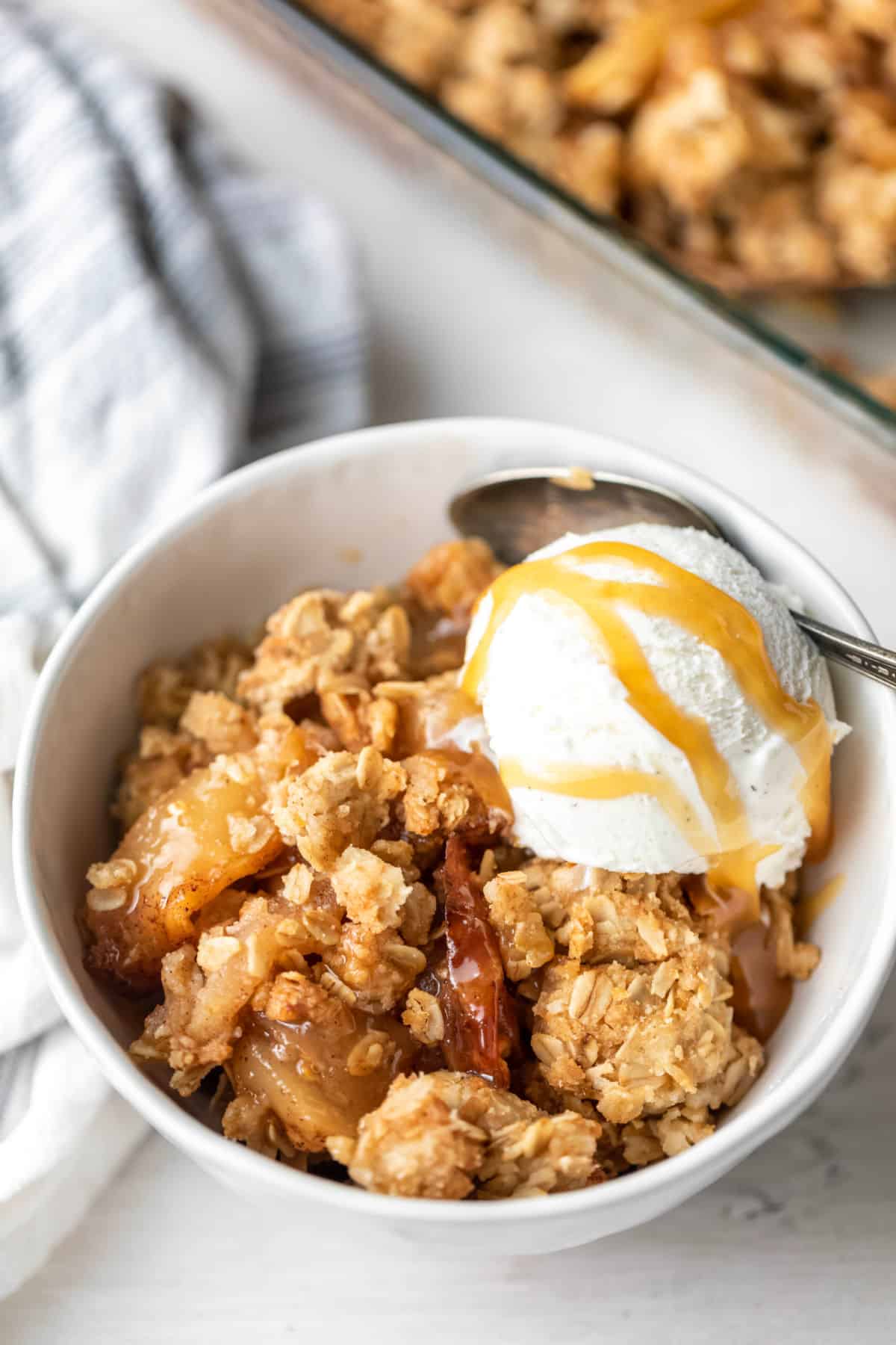 A white bowl of apple crisp with a scoop of vanilla ice cream and a spoon on it. 