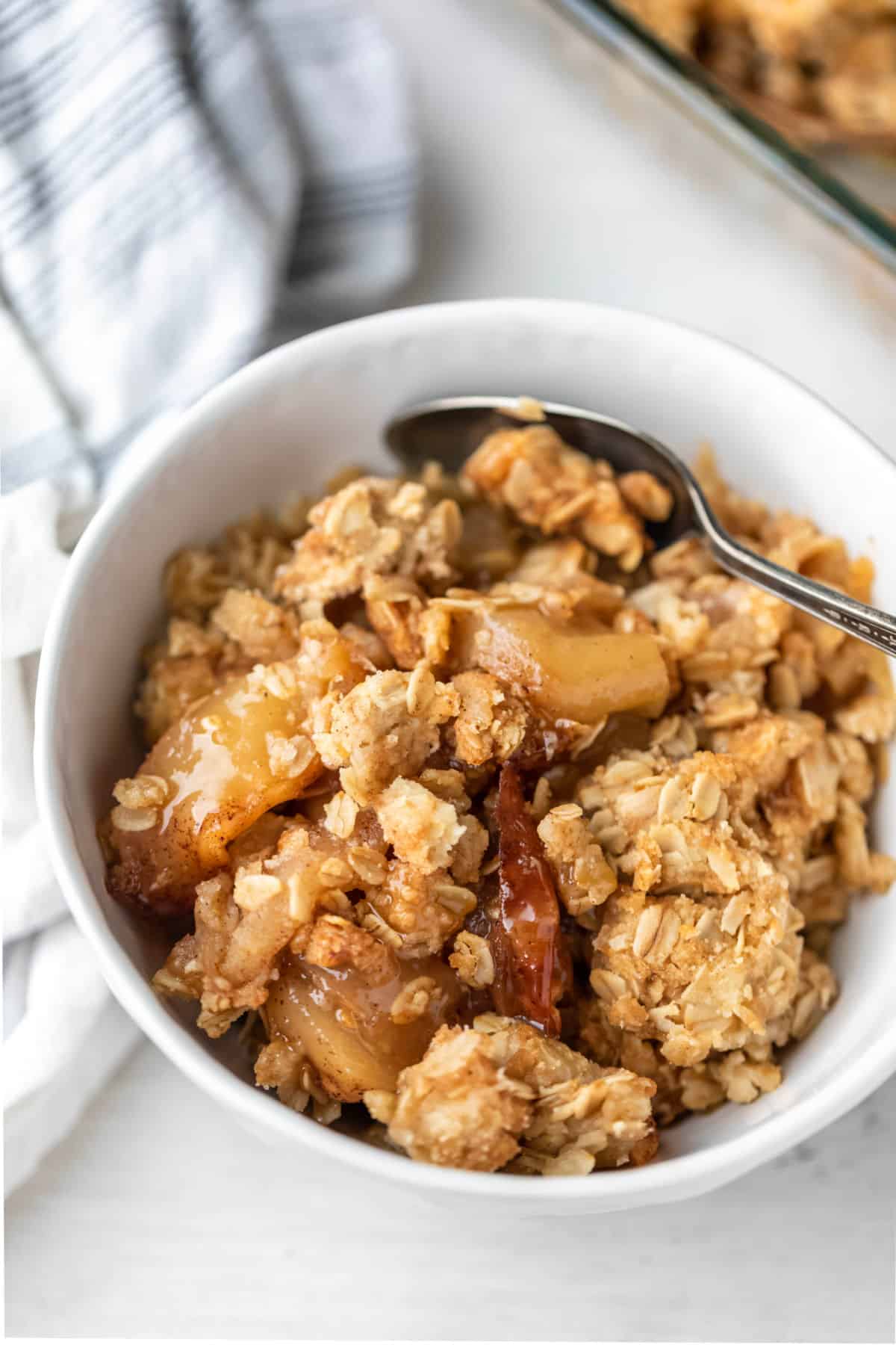 A bowl of apple crisp next to a linen and baking pan of apple crisp. 