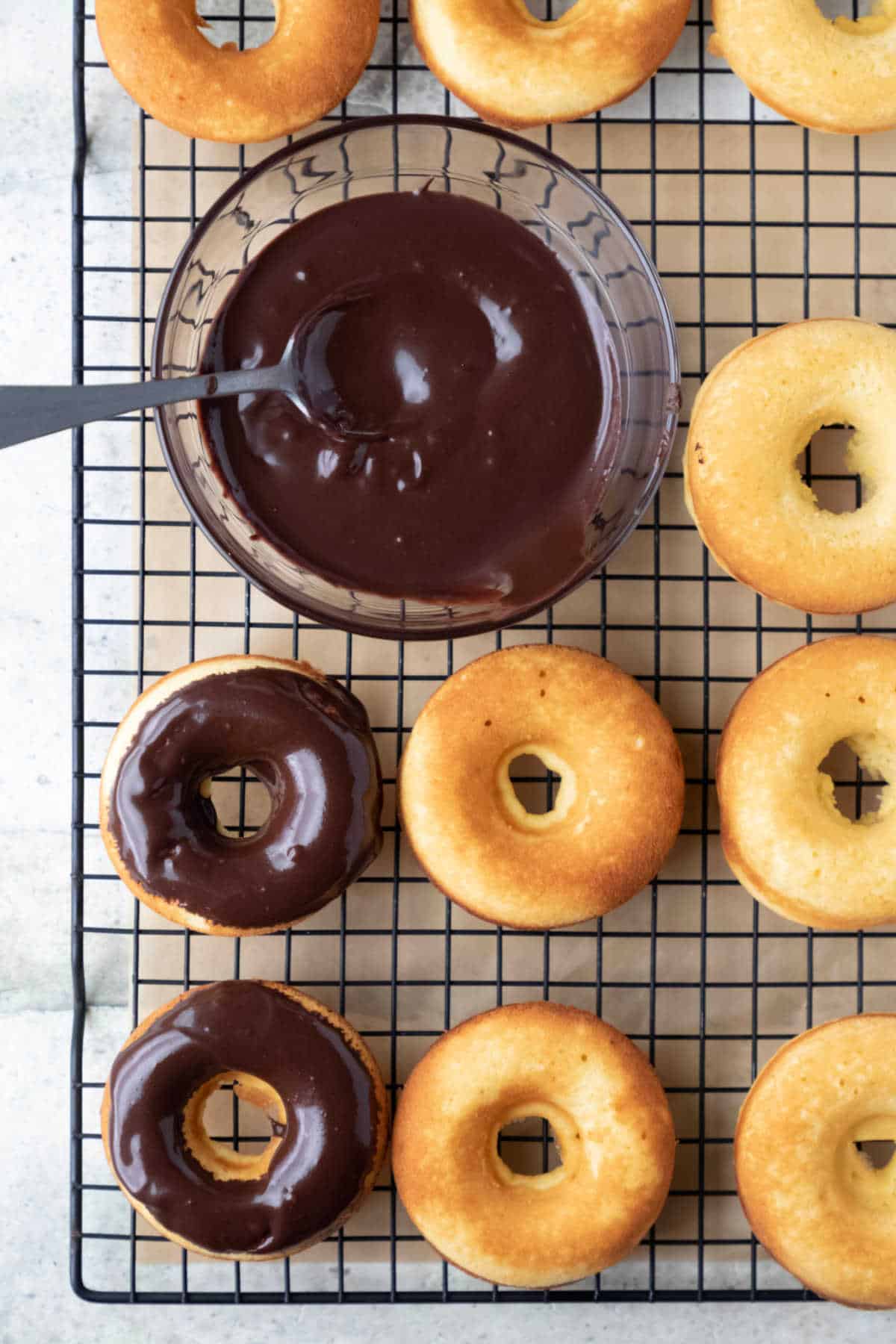 Two baked donuts topped with chocolate glaze next to a bowl of chocolate glaze. 