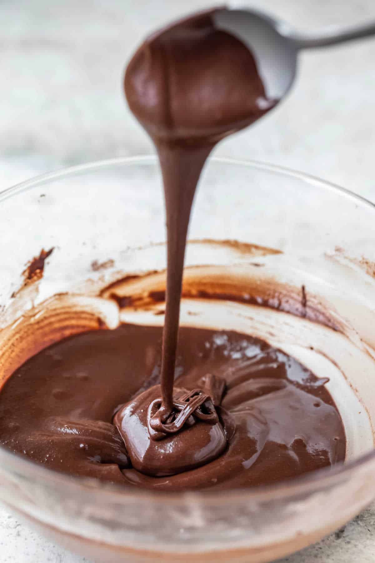 A spoon pouring chocolate glaze into a glass bowl.