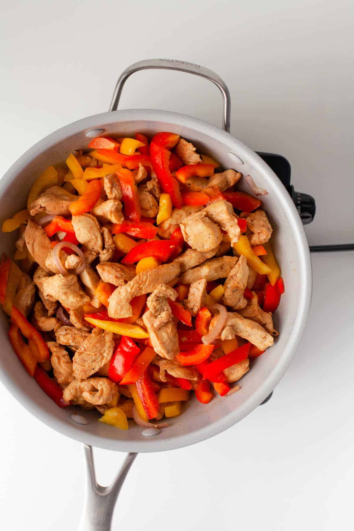 Chicken strips onion slices and slices of bell pepper cooking in a skillet. 