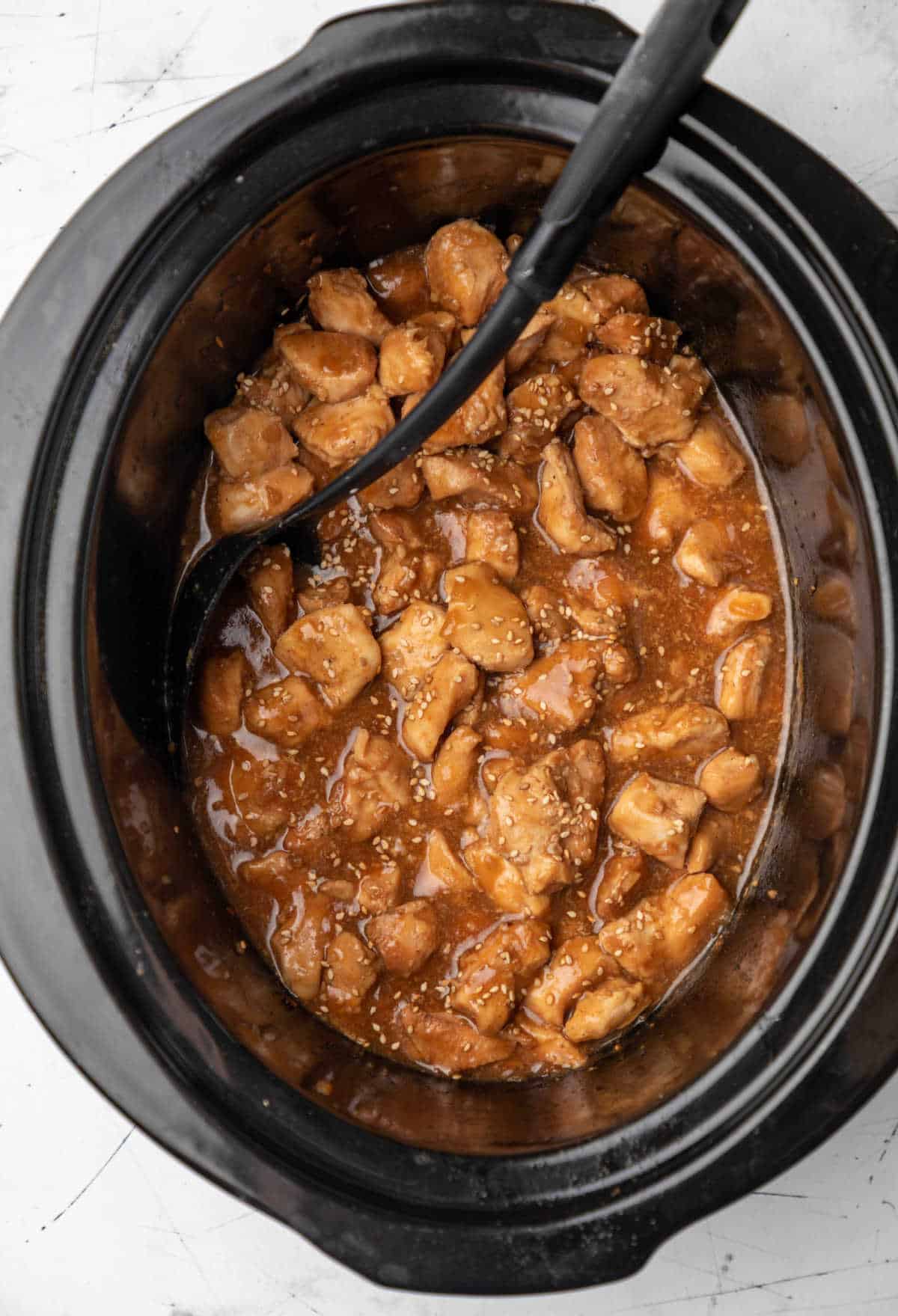 Sesame chicken in a crock pot with a black spoon in it. 