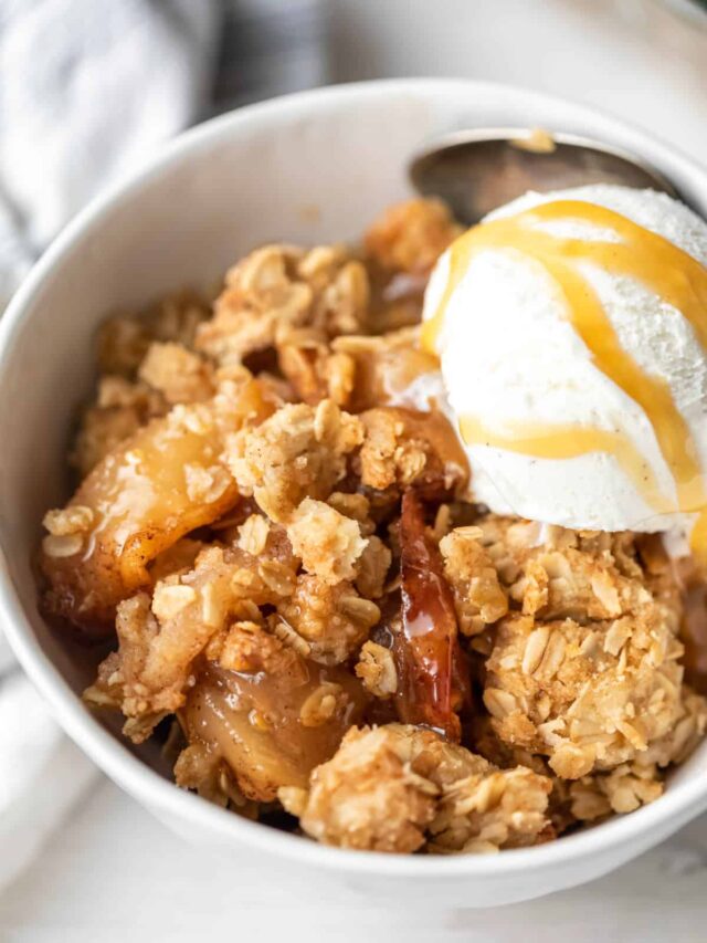 A white dish with apple crisp and a scoop of vanilla ice cream topped with caramel sauce.