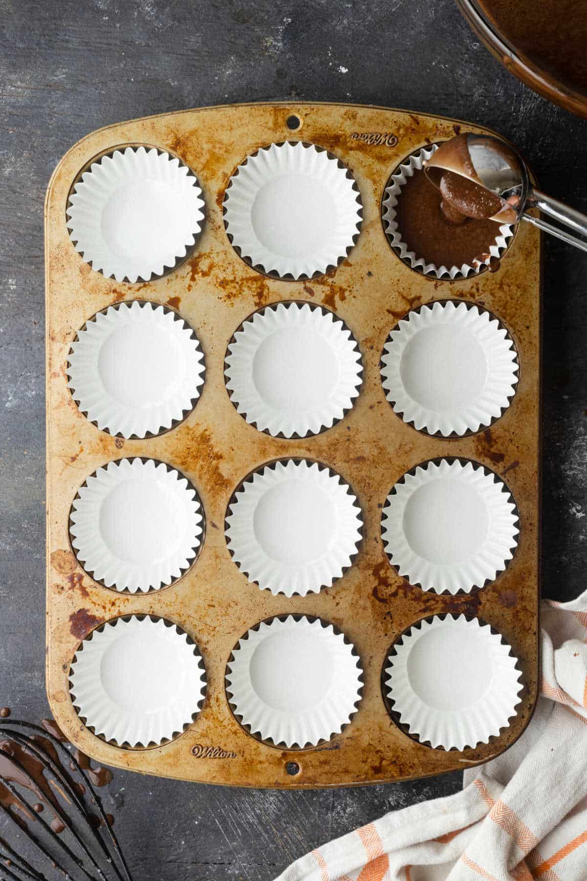 An ice cream scoop filling up a muffin pan with chocolate batter.