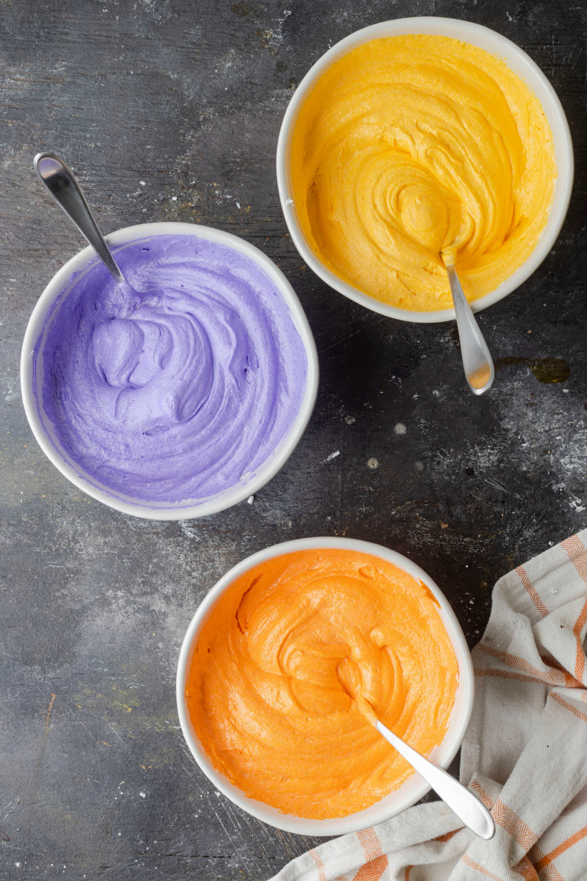 A mixing bowl of yellow frosting purple frosting and orange frosting. 