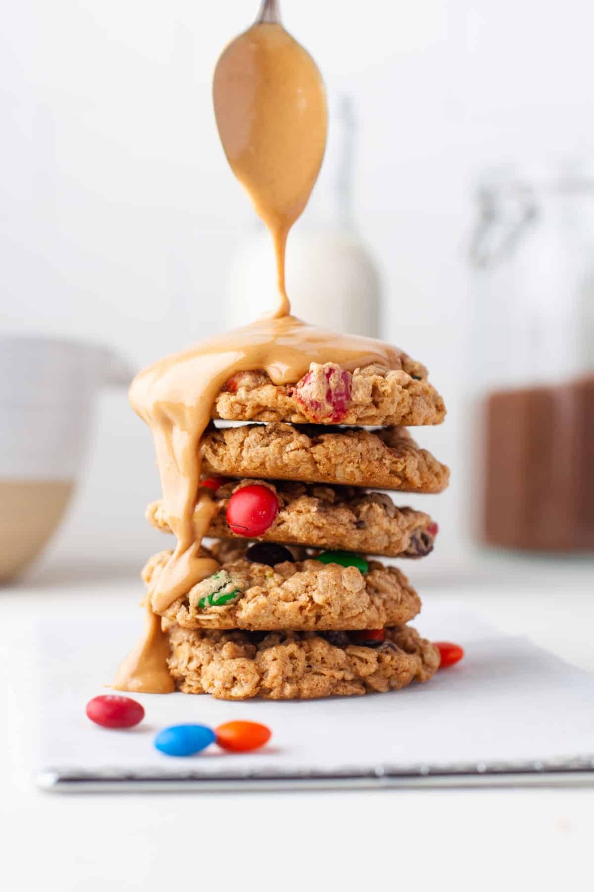 A spoon pouring peanut butter onto a stack of monster cookies. 
