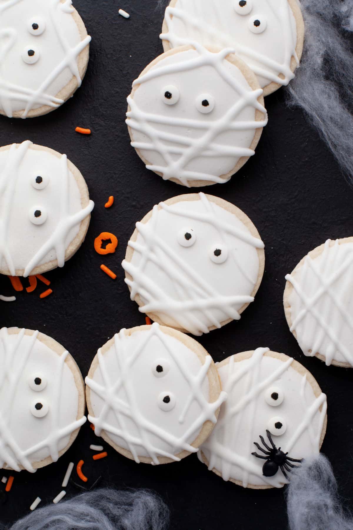 Mummy sugar cookies on a black background with plastic spiders and Halloween sprinkles around them. 