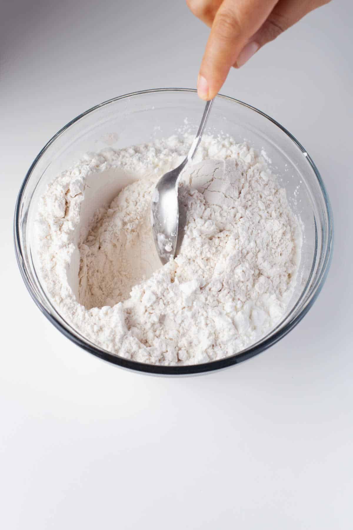 A spoon stirring flour cornstarch and salt together in a glass mixing bowl. 