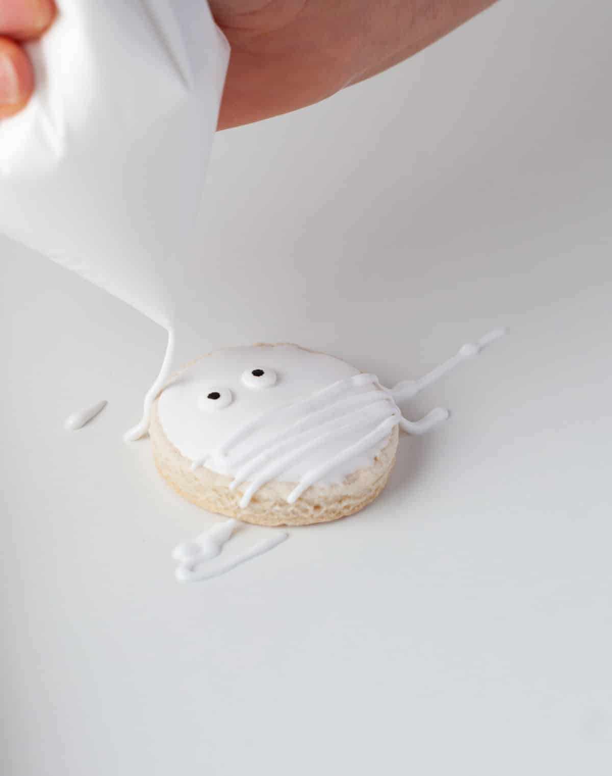 An icing bag drizzling royal icing onto a mummy sugar cookie. 