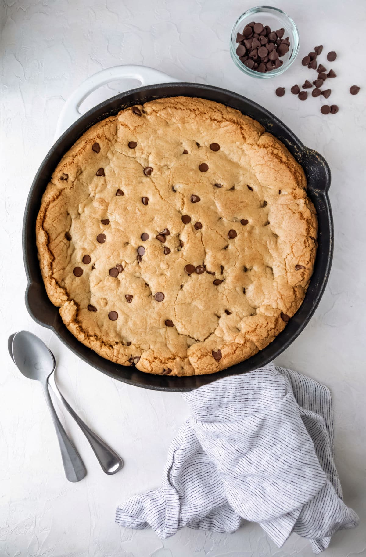 A pizookie next to two spoons and a bowl of chocolate chips. 