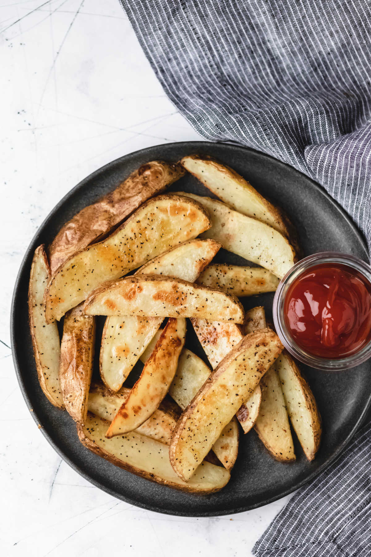 A plate of potato wedges and ketchup next to a gray striped linen. 