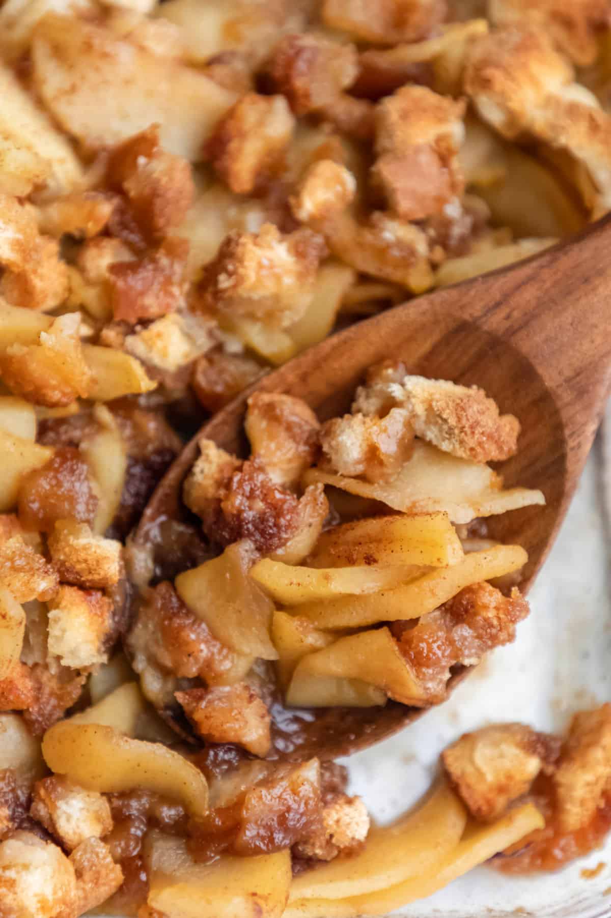 A wooden spoon scooping up apple brown betty from a baking dish.