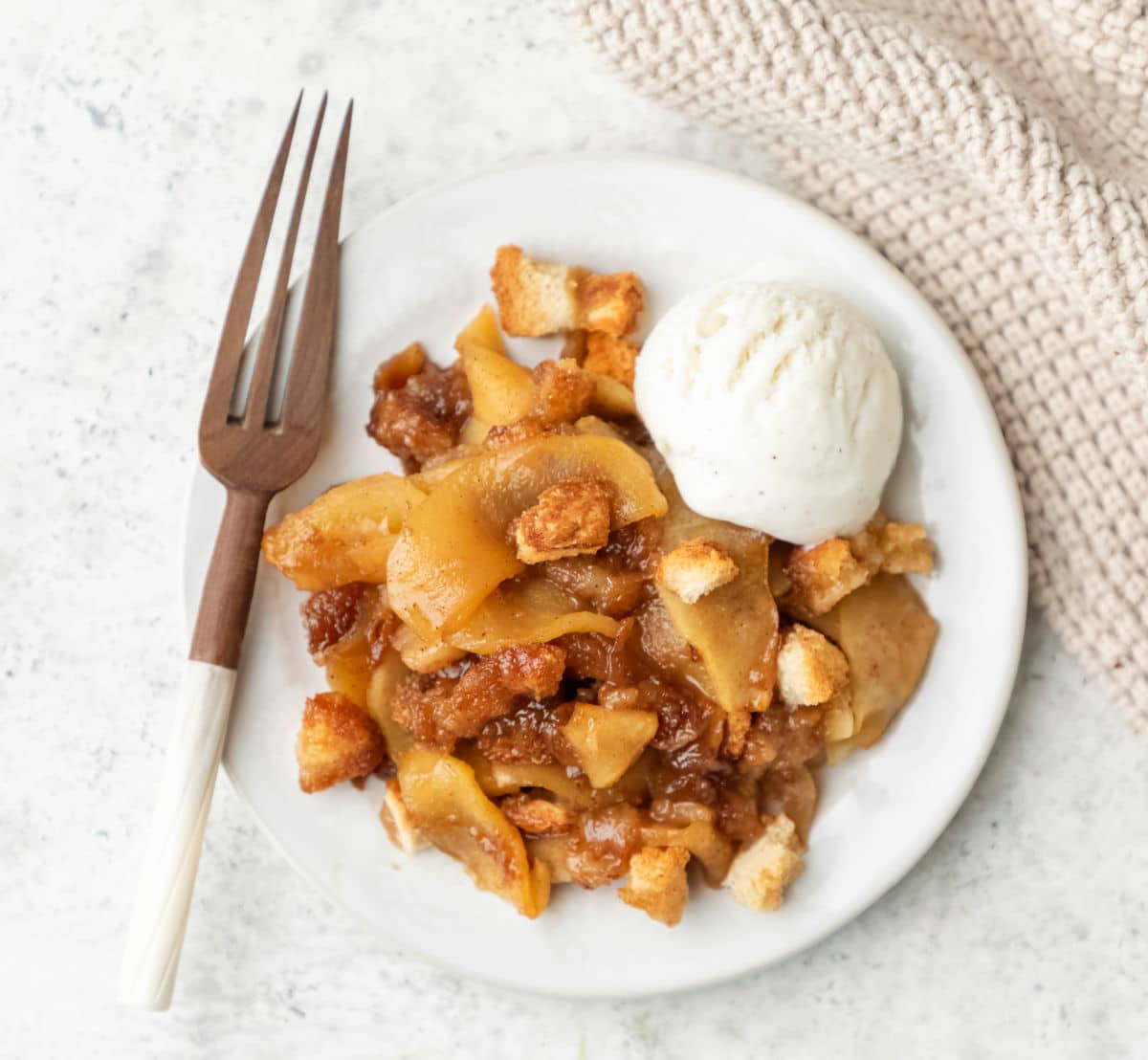 A white plate with apple brown betty a scoop of vanilla ice cream and a wooden fork on it.
