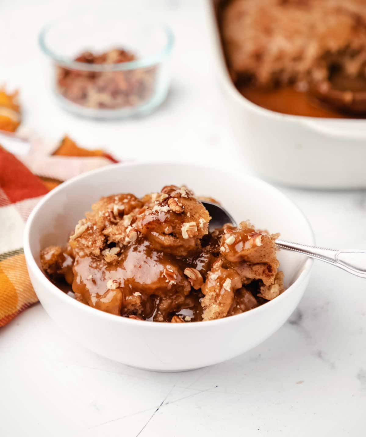 A white dish of caramel apple pudding cake behind a bowl of apple pudding cake. 