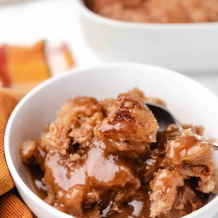 A white bowl of caramel apple cobbler cake next to an orange plaid napkin.