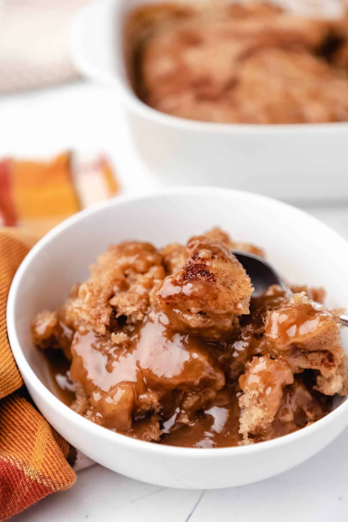 A white bowl of apple pudding cake next to an orange plaid napkin. 