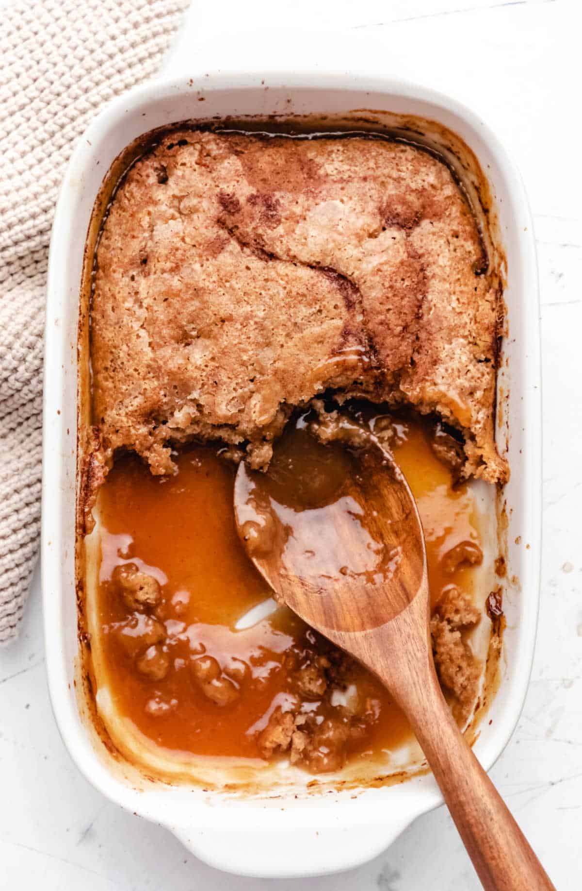 A wooden spoon in a partial dish of caramel apple cobbler cake. 