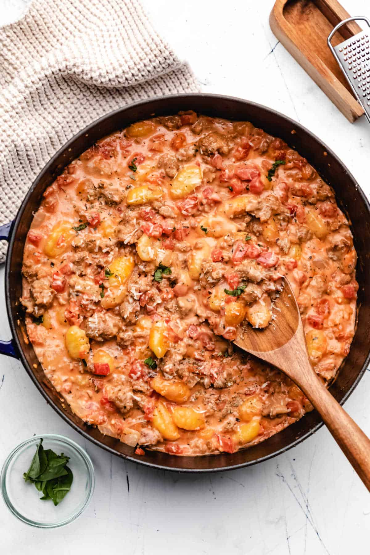 A pan of creamy sausage gnocchi next to a dish of basil. 