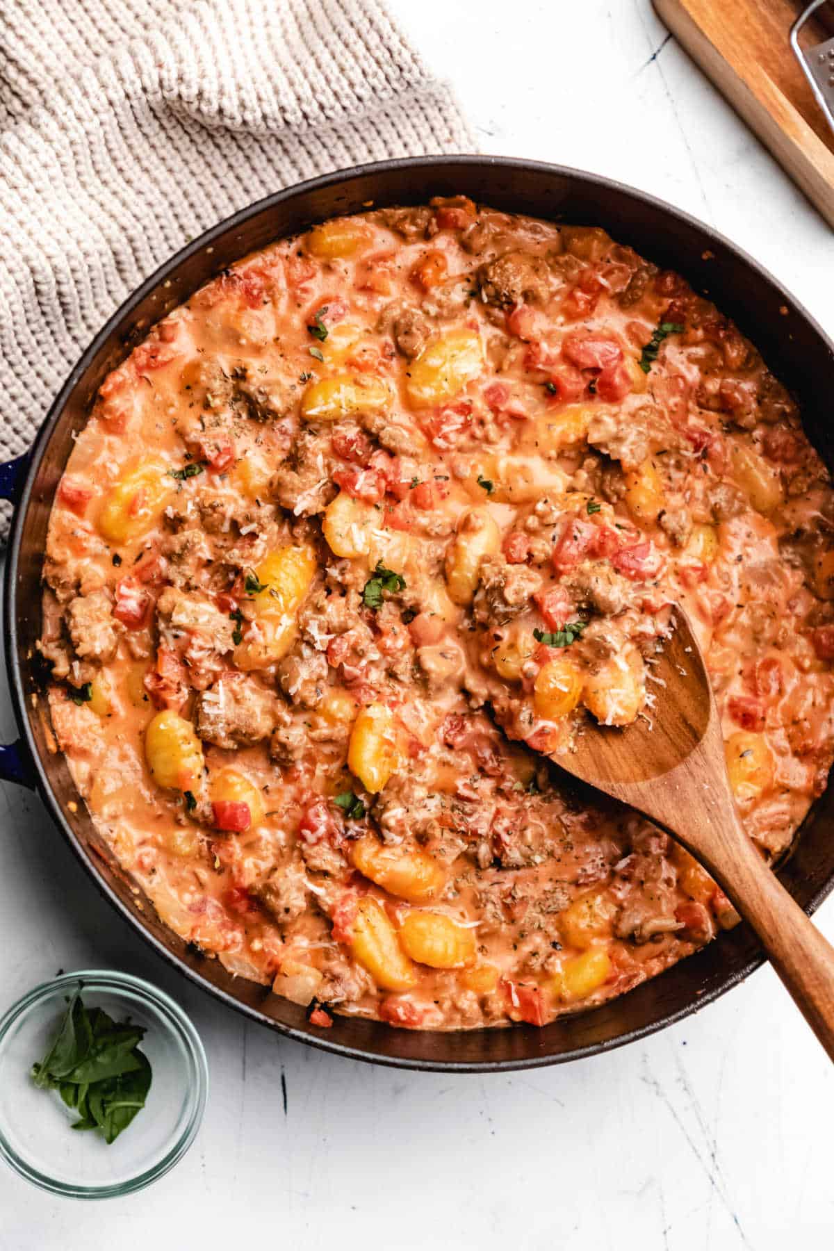 A skillet of creamy sausage gnocchi next to a chunky knit cloth. 